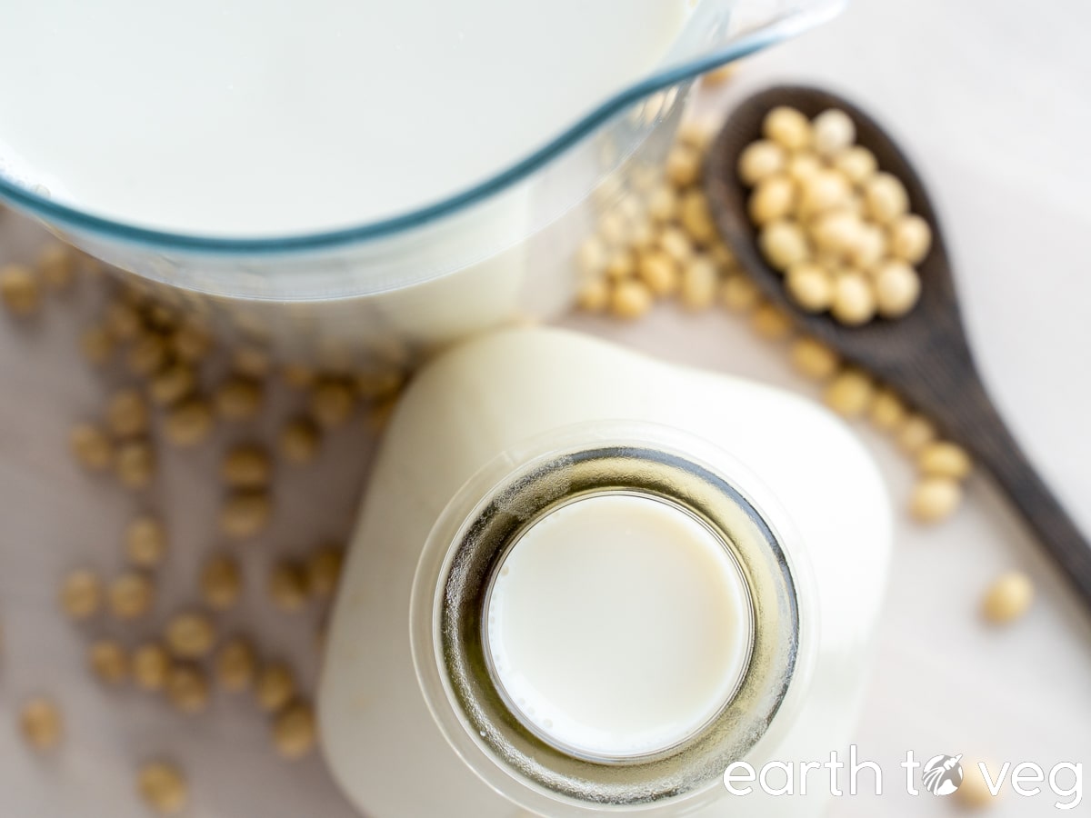 homemade soy milk in a glass jar next to a glass pitcher, with some soybeans on the tabletop.