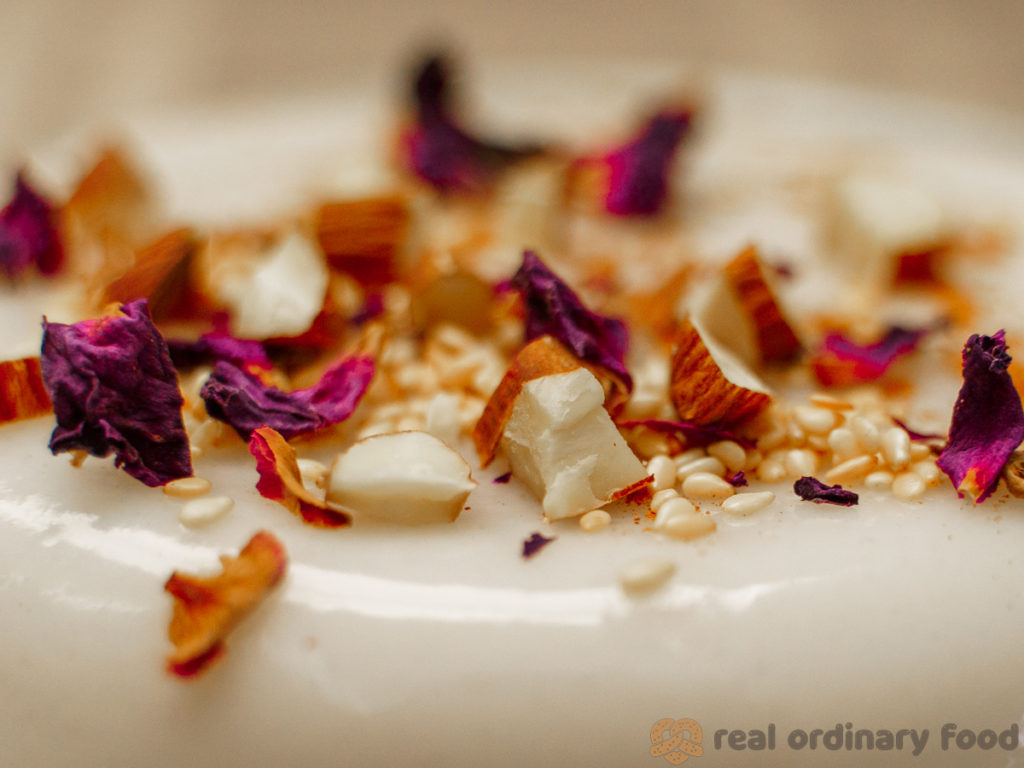 A close up shot of vegan muhallebi, decorated with rose petals, sesame seeds, and chopped almonds.