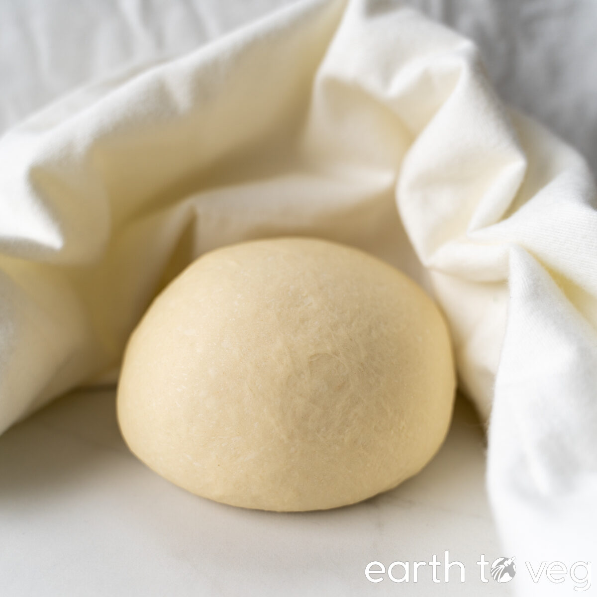A round, smooth ball of hot water dough on a white counter with a soft white cloth around it.