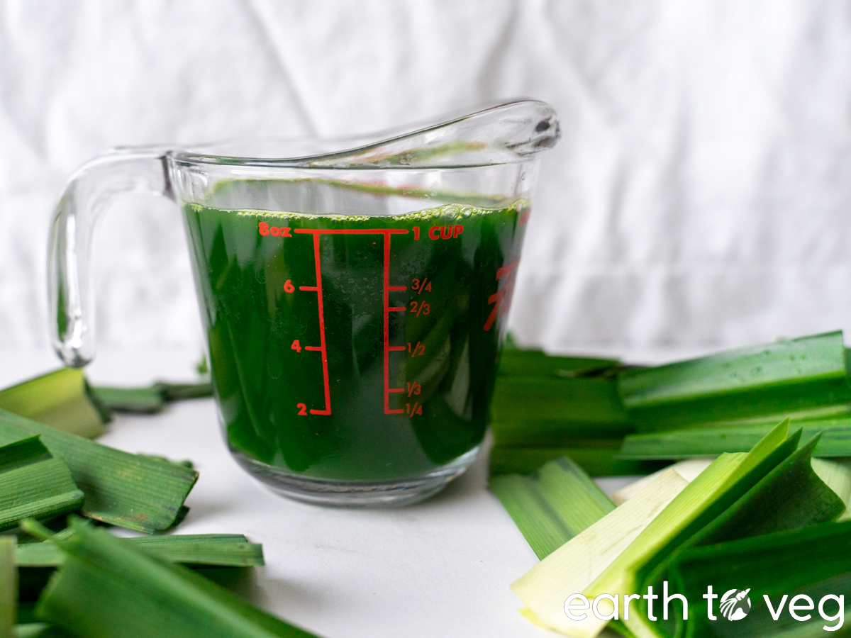 A glass measuring cup filled with fresh pandan juice sits on a white tabletop surrounded by chopped pandan leaves.