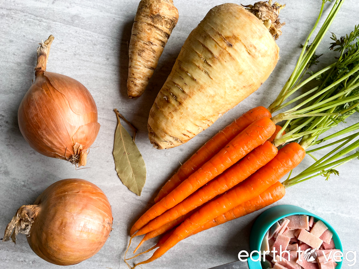 Onions, bay leaf, parsnips, carrots, and vegan bacon are laid out on a grey counter.