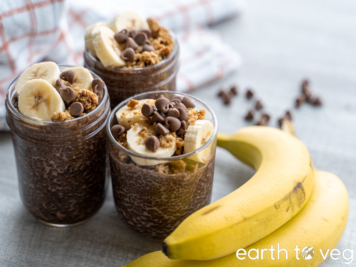 three jars of banana chocolate chia pudding next to a bunch of bananas on a gray counter.