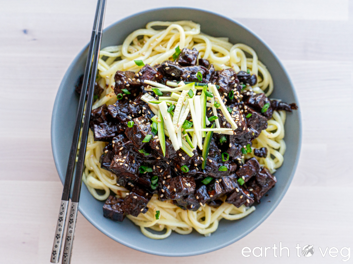 Vegan jjajangmyeon in a bowl with chopsticks on the side, topped with julienned cucumber and sesame seeds.
