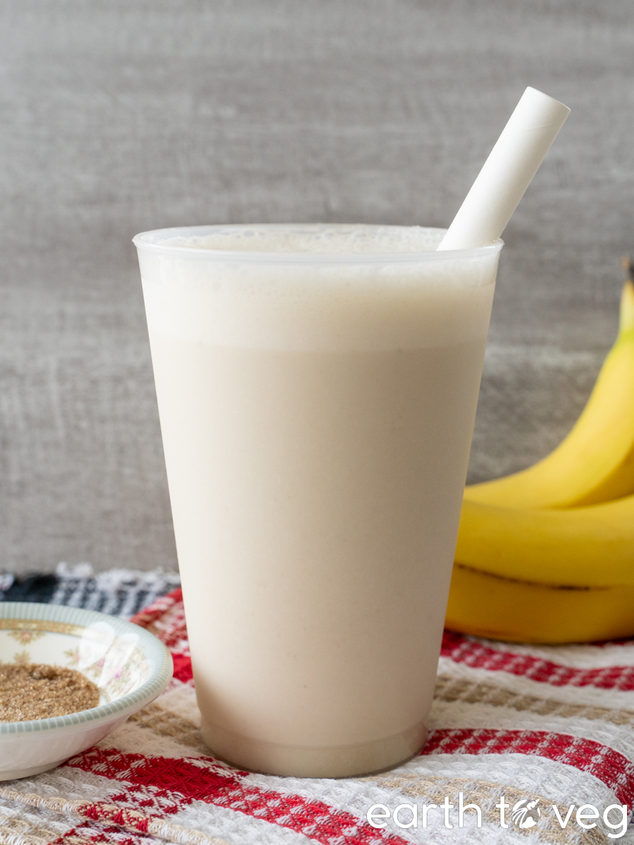 A banana smoothie cup with a white paper straw is on a red and black waffle-patterned towel, in front of some bananas and next to a small bowl of sugar.
