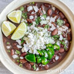 A pot of frijoles de la olla topped with cilantro, jalapeno slices, cheese, and sliced lime wedges.