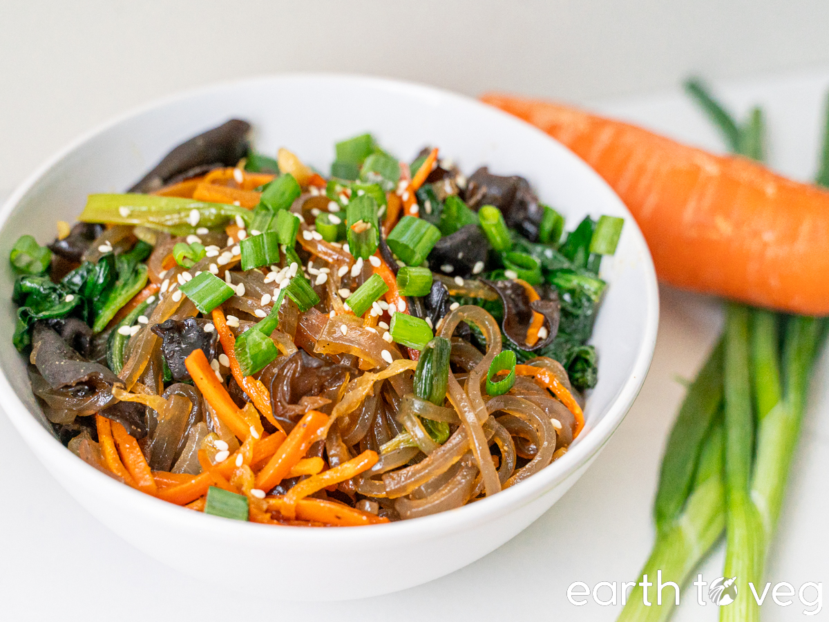 A bowl of japchae sits on a white table next to fresh scallions and a carrot.