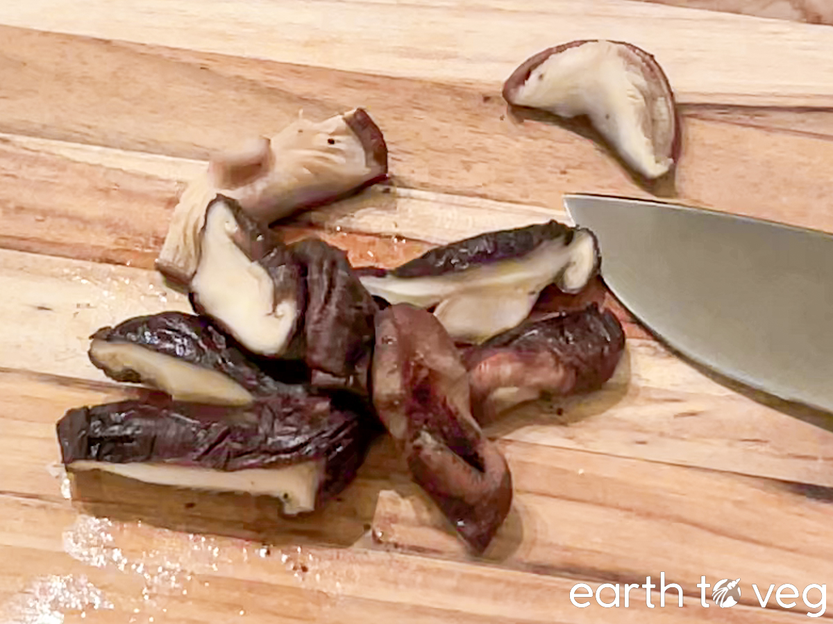 Soaked shiitake mushrooms are sliced on a teak cutting board.