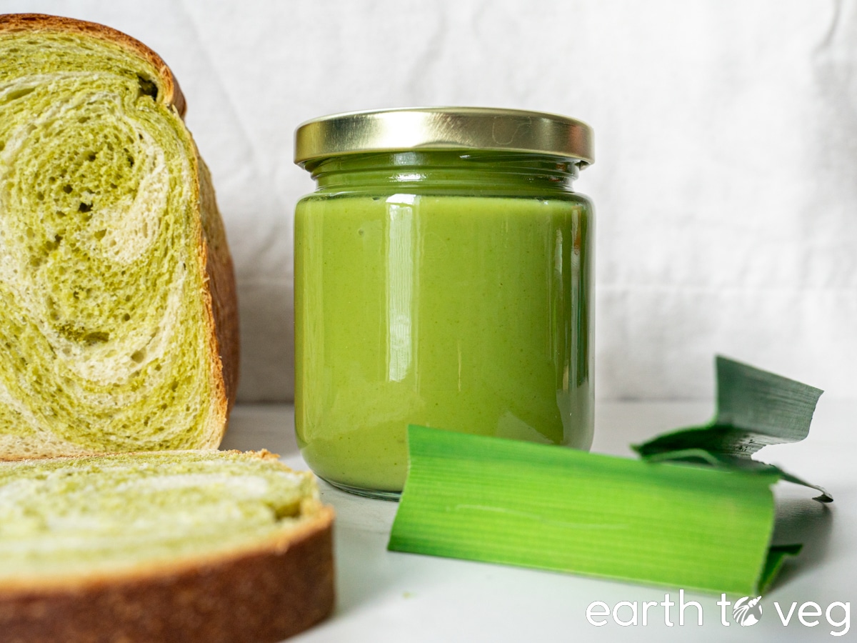 A jar of vegan pandan kaya sits on a white table next to fresh pandan leaves and a loaf of pandan bread.