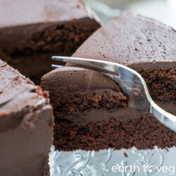 A metal fork cuts into a double-layer chocolate cake with a thick layer of chocolate frosting.
