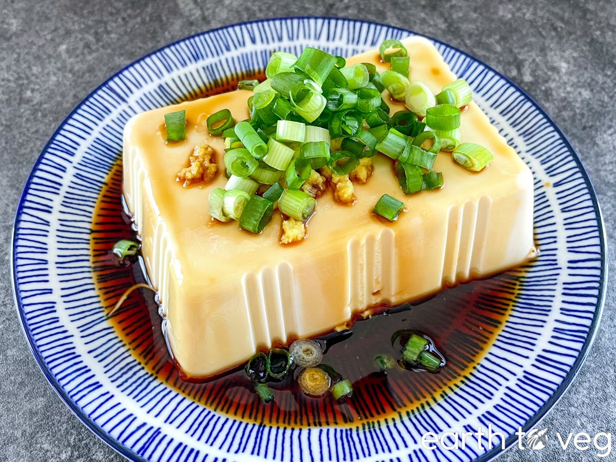 A block of cold silken tofu topped with fresh ginger, scallions, and Japanese soy sauce on a blue china plate.