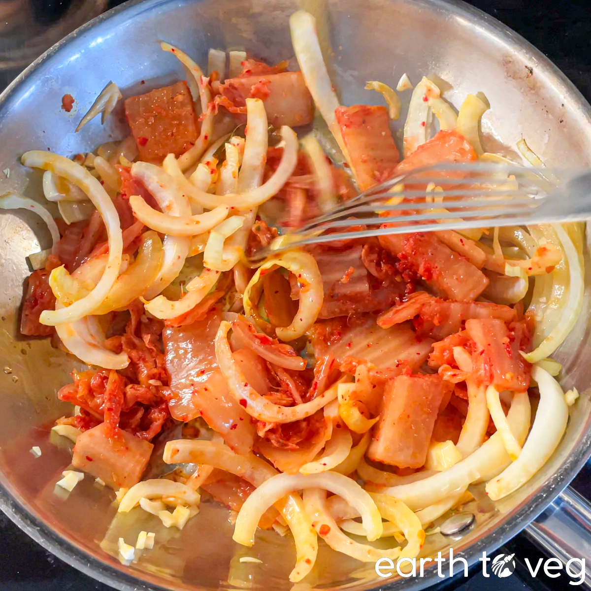 Kimchi, onions, and garlic are sautéed together in a stainless steel pan.