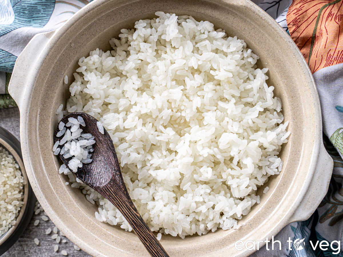 A wooden spoon rests on a clay pot full of cooked white rice next to some colourful tablecloths.