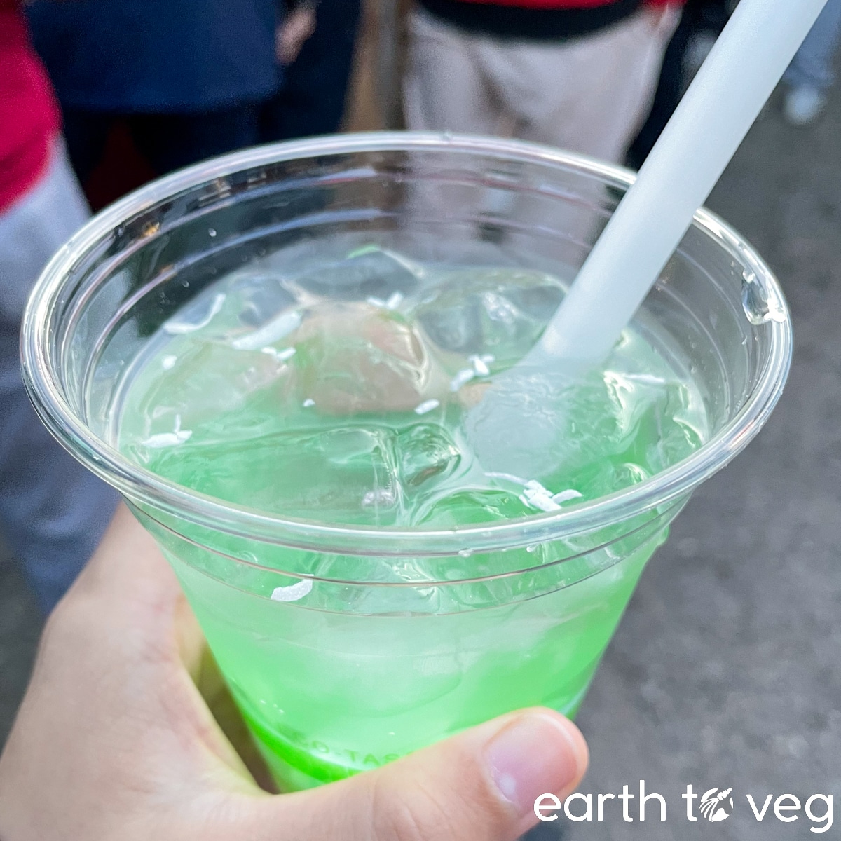 Hand holding up a small plastic cup of pandan coconut lychee drink filled with ice cubes.