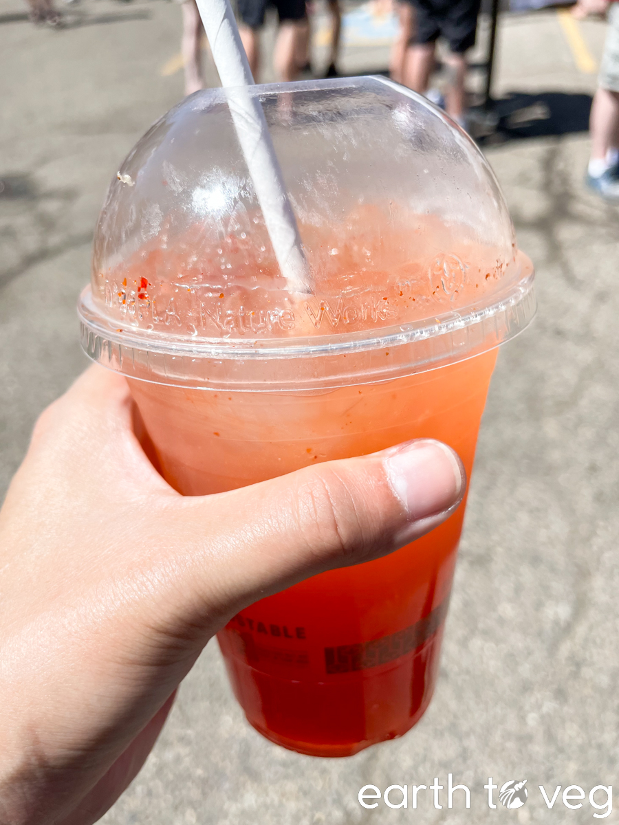 A tall plastic cup filled with orange passion fruit tea is held over the concrete midway.
