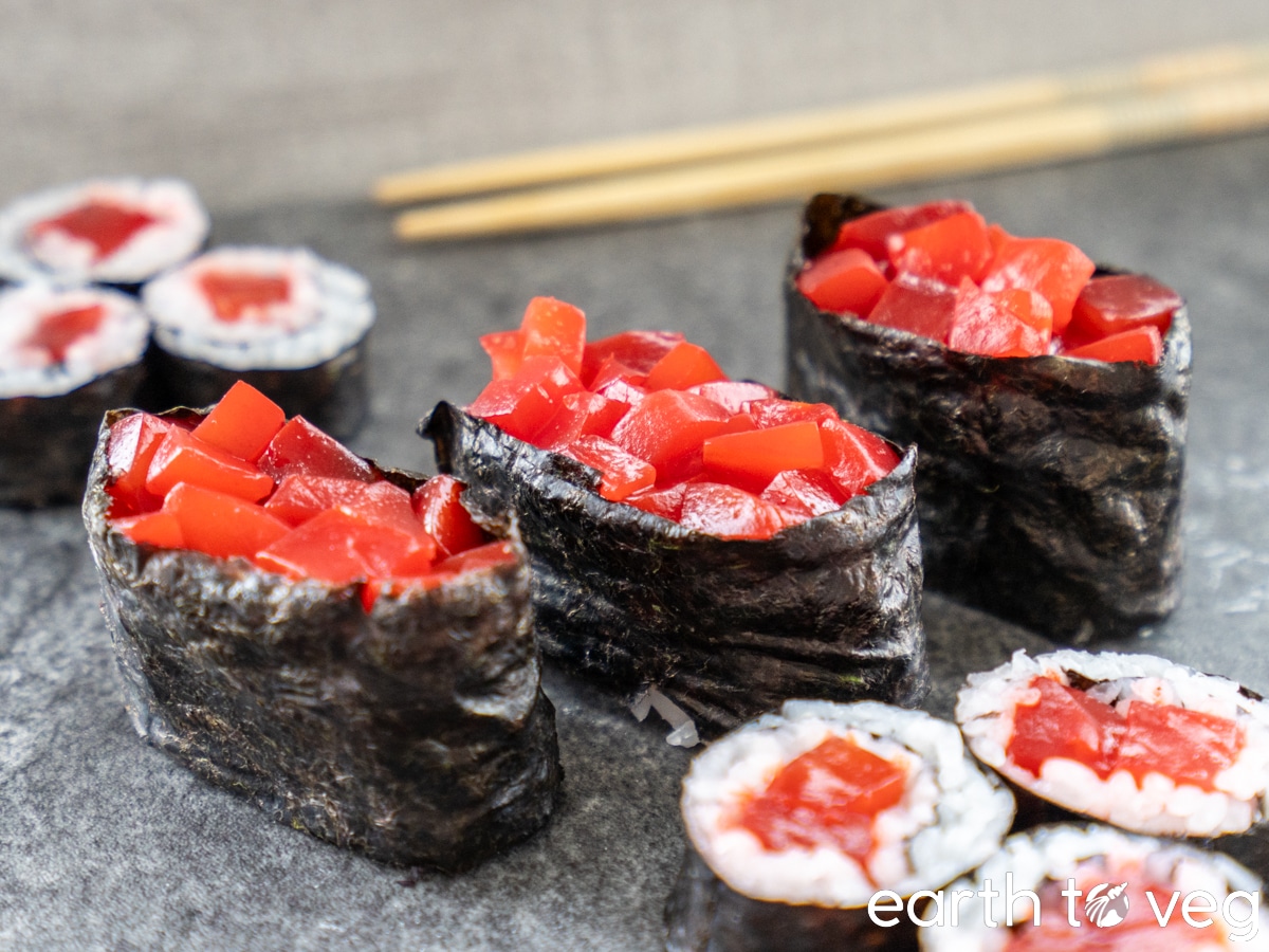 Three pieces of vegan gunkan maki are flanked by vegan maki rolls on a grey surface, with chopsticks in the background.