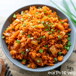 A grey bowl full of kimchi fried rice sprinkled with diced scallions set on a newspaper clipping, with fresh whole scallions in the background.