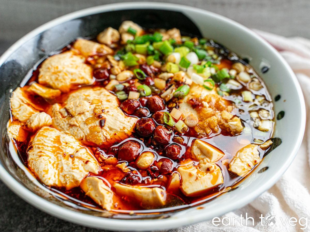 Savoury doufunao in a large bowl covered with soy sauce, chili crisp, scallions, and toasted peanuts.