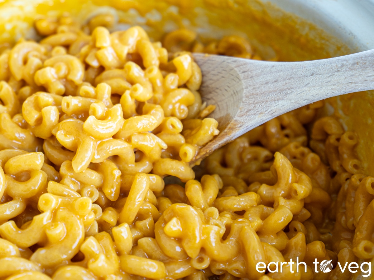 A wooden spoon ladles a big pile of vegan macaroni and cheese from a Dutch oven.