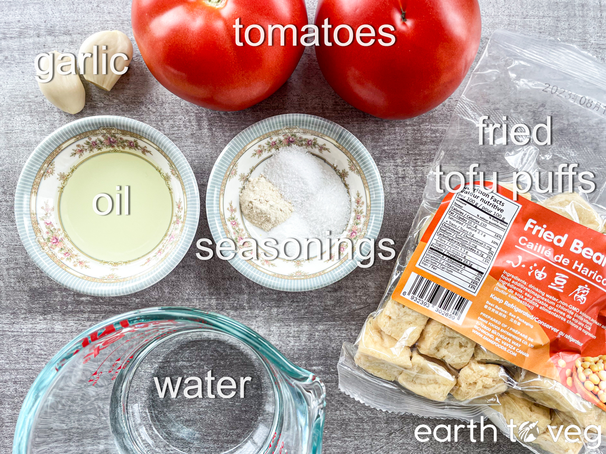 Ingredients for Vietnamese Tomato Tofu, such as tomatoes, tofu puff, oil, and seasonings, are laid out on a grey surface.