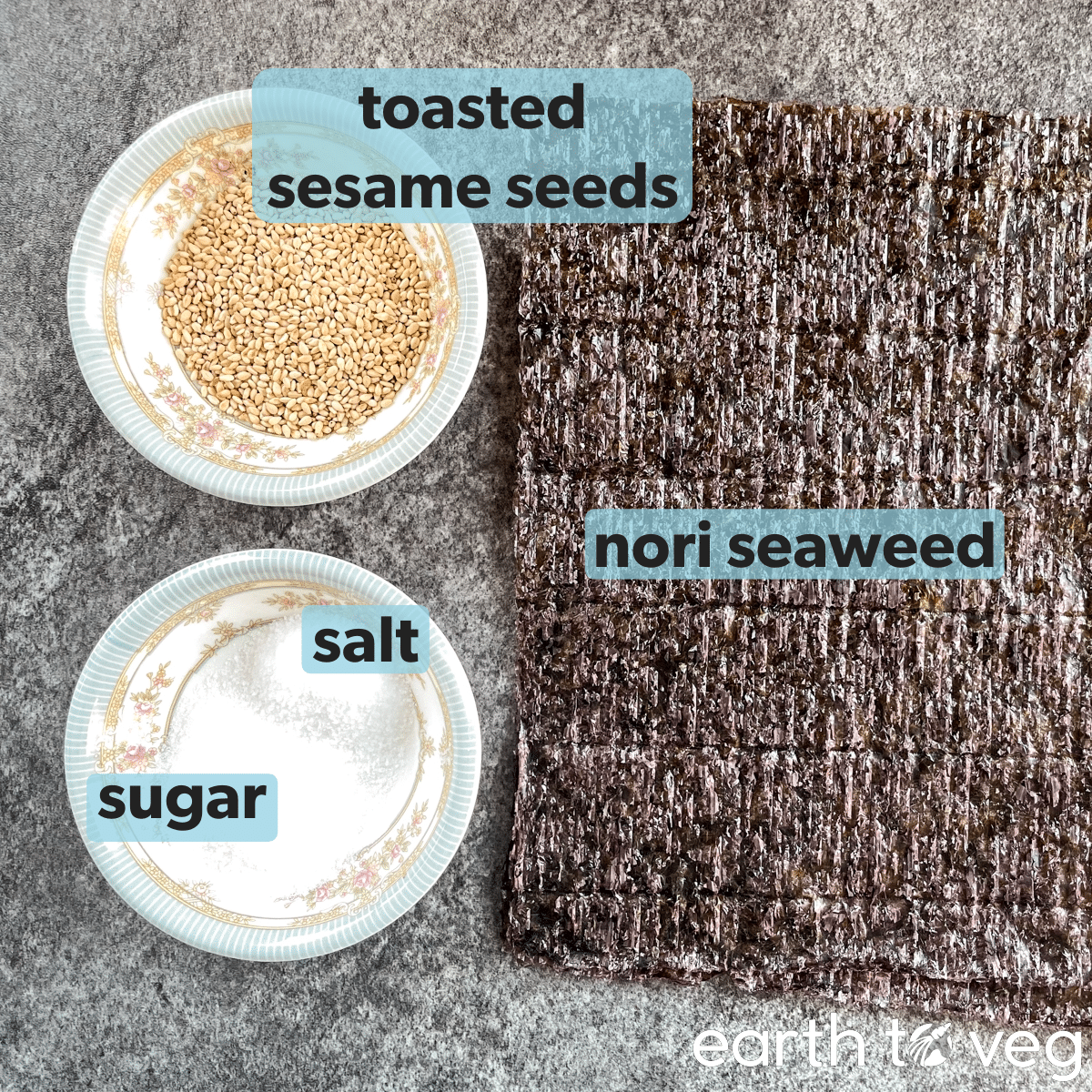 Toasted sesame seeds, salt, sugar, and nori sheets laid out on a grey countertop.