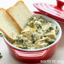 A small red cocotte of vegan spinach artichoke dip with two slices of Melba toast dipped into it.