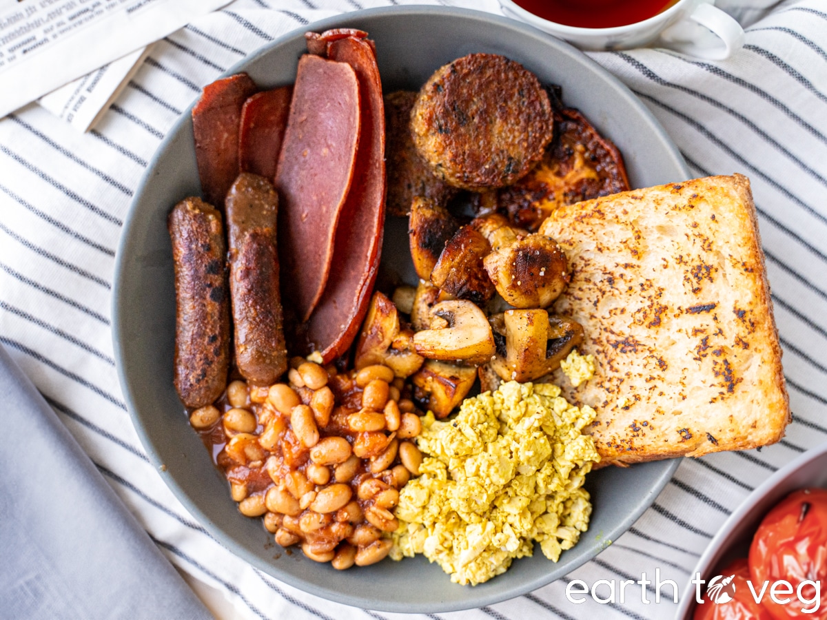 A vegan full English breakfast spread on a grey plate with stewed tomatoes and hot black tea on the side.