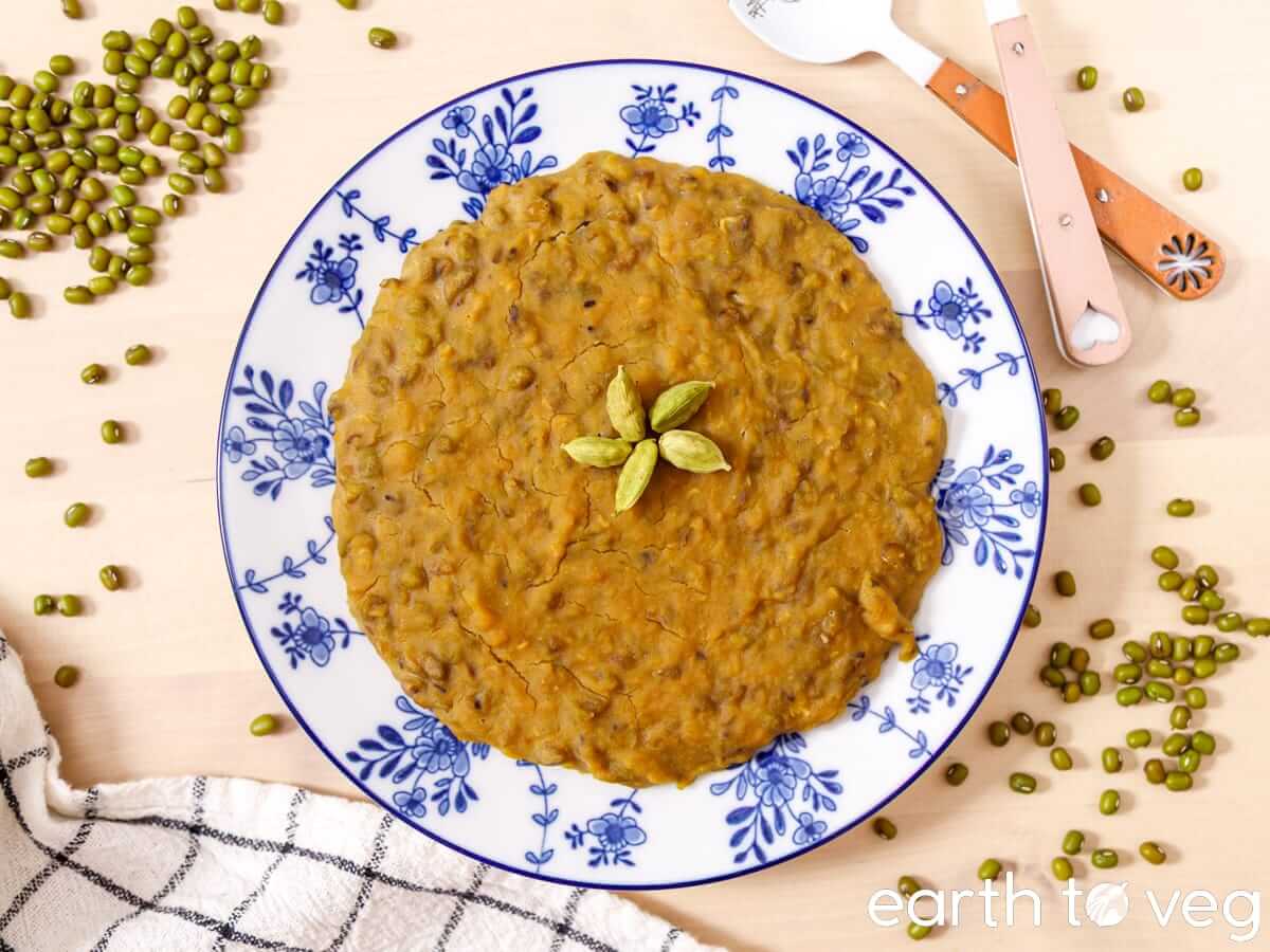 Plate of godshem topped with cardamom pods, framed by scattered raw mung beans and a checkered tea towel.