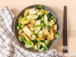 A bowl of bok choy tofu stir fry with marinated tofu planks and tender baby bok choy, set on a wooden table with black chopsticks and a checkered linen napkin.