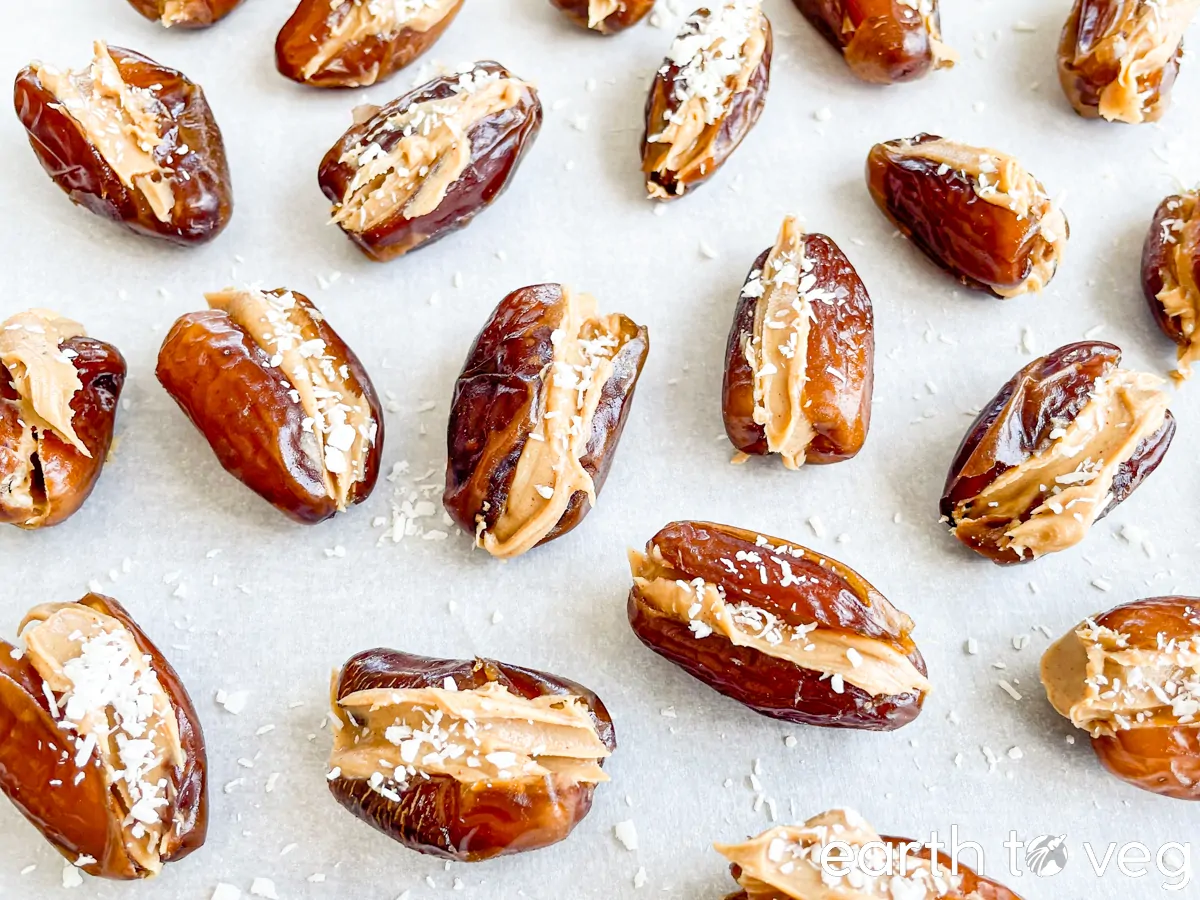 A close-up of halved dates on a parchment-lined surface, each stuffed with peanut butter and sprinkled with shredded coconut, arranged in a grid-like pattern.