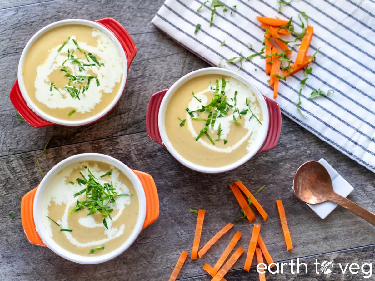 Three bowls of creamy soup garnished with fresh herbs sit on a wooden surface. Two bowls have red handles, and one has orange. Carrot sticks and a wooden spoon lie nearby on a striped cloth. The words "earth to veg" are written in the bottom right corner.